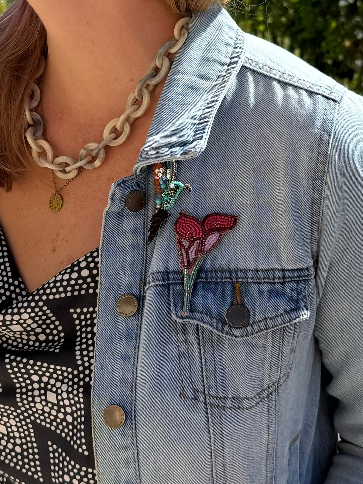 A close-up of a person wearing a light-wash denim jacket over a black and white patterned top, accessorized with a chunky chain necklace and a colorful embroidered floral pin on the pocket.